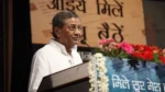 Ghanshyam Tiwari speaking at a public event, standing at a podium with flowers and a Hindi backdrop.
